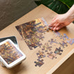 Person assembling Cat City Life Jigsaw Puzzle in Tin, featuring cats in an urban scene, with puzzle pieces on a wooden table.