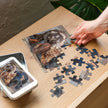 Dachshund Duo Jigsaw Puzzle in Tin showing two dachshunds with dapple and black-and-tan coats, near a white tin on a wooden table.